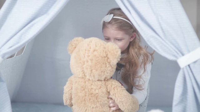 Little cute girl with blond hair sitting in a toy house surrounded by soft toys. Girl playing with a toy hare and a soft teddy bear. Wigwam for children in a room.