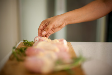 African American woman reaches for a piece of chicken, her hand in focus. In the foreground  blurred shot of drumsticks on a cutting board with rosemary and thyme leaves. .