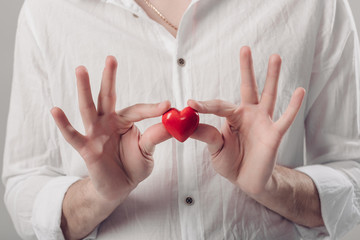 man in white shirt holds in hands red heart on gray background. 