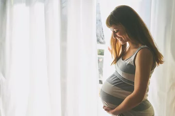 Poster Young beautiful pregnant woman standing near window at home © SianStock
