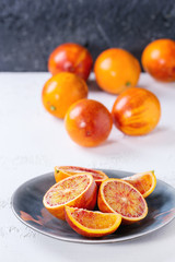 Sliced and whole ripe juicy Sicilian Blood oranges fruits on ceramic plate over white and gray concrete texture background.
