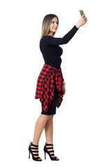 Pretty young stylish woman in black dress taking selfie photo with mobile phone. Full body length isolated over white studio background. 