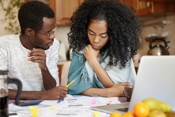 Stressed young African family of two looking desperate, sitting at table with lots of papers and laptop, trying to support each other, have to move out of their apartment because of non-payment