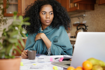 Fototapeta na wymiar Worried beautiful African-American woman drinking coffee at kitchen table, planning budget, reviewing finances, calculating expenses and paying utility bills online, sitting in front of open laptop
