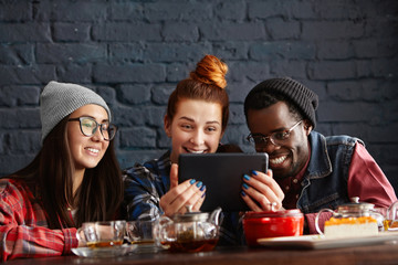 Three students enjoying free wi-fi, using digital tablet at cafe during lunch break. Multiethnic group of young happy people having fun at restaurant, talking and laughing while surfing Internet