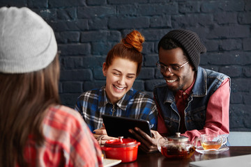 People, leisure, communication and modern technology concept. Happy interracial couple enjoying high-speed connection at restaurant during lunch, browsing internet on digital tablet and laughing