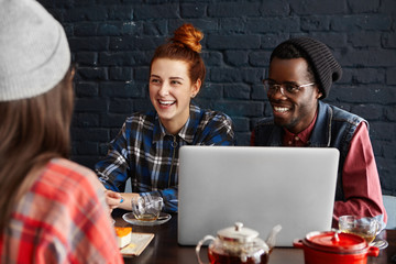Happy Caucasian redhead girl and her dark-skinned boyfriend wearing glasses and stylish hat sitting at cafe in front of open laptop, laughing and looking at their common female friend in foreground