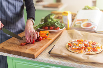 Close-up view of young woman s hands cutting vegetables on board for pizza according to recipe book