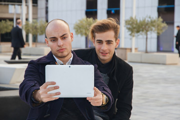 Two young business people doing a selfie on a break from work with a tablet. They are sitting on a bench outside the office. 