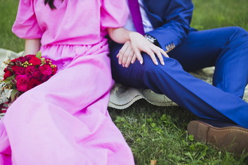 Happy couple in love sitting on green grass on their wedding day. Bride in simple stylish pink dress holds beautiful wedding bouquet, nice flowers. Holding hands. Happy people on summer day outdoors