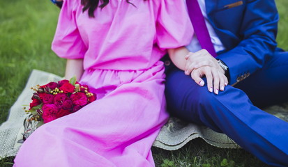 Happy couple in love sitting on green grass on their wedding day. Bride in simple stylish pink dress holds beautiful wedding bouquet, nice flowers. Holding hands. Happy people on summer day outdoors