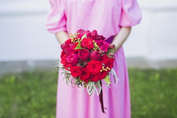 Happy bride in a simple pink wedding dress holding a beautiful bouquet of red roses, peonies and green leaves. Woman in a stylish dress celebrating summer day wedding, blurred background.