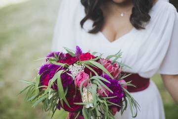 Happy bride in a simple white wedding dress holding a beautiful bouquet of flowers and green leaves. Woman in a stylish dress celebrating summer day wedding, blurred background.