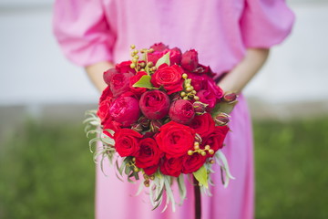 Happy bride in a simple pink wedding dress holding a beautiful bouquet of red roses, peonies and green leaves. Woman in a stylish dress celebrating summer day wedding, blurred background.