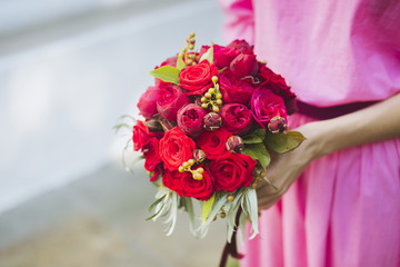 Happy bride in a simple pink wedding dress holding a beautiful bouquet of red roses, peonies and green leaves. Woman in a stylish dress celebrating summer day wedding, copy space, blurred background