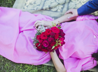 Happy couple in love lying in green grass on their wedding day. Bride in simple stylish pink dress holds beautiful wedding bouquet, nice flowers. Happy people on summer day outdoors.