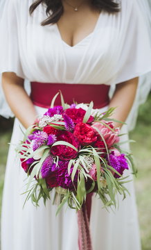 Happy bride in a simple white wedding dress holding a beautiful bouquet of flowers and green leaves. Woman in a stylish dress celebrating summer day wedding, blurred background