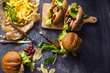 Fast food on the table: hot dog sausage and vegetables, burger with pork or beef and French fries with tomato sauce, basil leaves