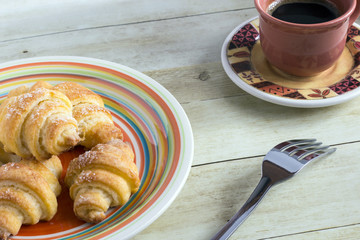 Cinnamon crescents in colored plate, a cup of coffee and fork on light wood background
