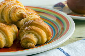 Cinnamon crescents in colored plate, a cup of coffee and cloth napkin on light wood background