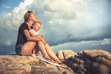 Loving young mother embrace her baby sitting on rocks with dramatic sky and clouds on background during sunset with copy space