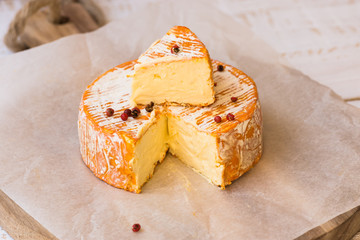 Top view of cut slice of French or German soft cheese with orange rind with mold, creamy texture, red pepper corns, waxed paper, cutting board