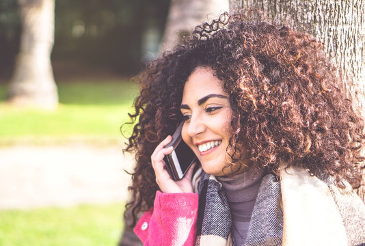 Portrait Of Pretty Young Woman Having A Phone Call Conversation With Mobile Smartphone While Standing Relaxed In Peaceful Place - Attractive Female Brunette With Curly Hair Talking On Cell Telephone