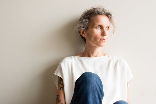 Middle Aged Woman In Cream Top And Jeans Sitting On Floor Looking Pensive Against Neutral Background (cropped And Selective Focus)