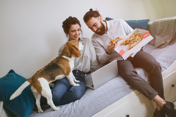 Young hipster couple home eating pizza with laptop and dog