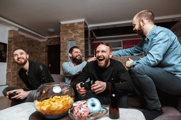 Men with a beard sitting on the couch at home talking and laughing with beer and joysticks in the hands of playing computer video games, one of them is eating a donut. 
