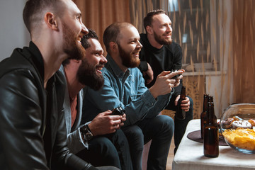 Men with a beard sitting on the couch at home talking and laughing with beer and joysticks in the hands of playing computer video games, one of them is eating a donut. 