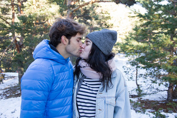 Fototapeta premium Young couple, boyfriend and girlfriend kissing and hugging in the mountains on a snowy day. They are on a date traveling to the snowy forest. 