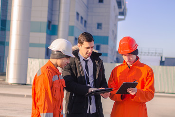 boss or Chief  instructs young team of  young engineers with a construction project on tablet. They wear overalls and safety helmets. Business modern background