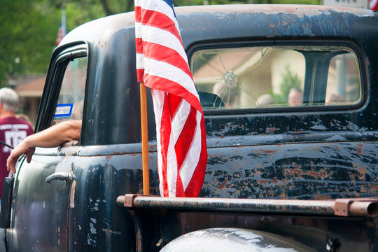 An American flag is attached to the back of an old, beat up pickup truck driving in an Independence Day Parade.
