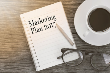 Concept  Marketing Plan 2017 message on notebook with glasses, pencil and coffee cup on wooden table.