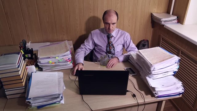 Office worker sits at a table in office, he works on a laptop, large piles of documents on the table

