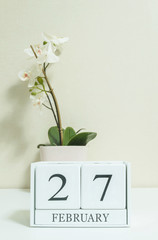 Closeup white wooden calendar with black 27 february word with white orchid flower on white wood desk and cream color wallpaper in room textured background in selective focus at the calendar