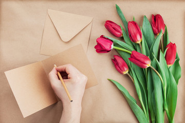 Spring tulip flowers and man hand signing a card on brown background top view in flat lay style. Greeting for Birthday, Womans or Mothers Day.