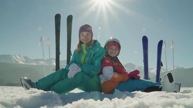 Mother and child girl are skiers sitting on snow against mountains. Family smiling and looking at camera