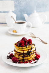 Stack of fried pancakes on a white plate. Chocolate, berries, raspberries, cranberries and blackberries on top. Gray concrete table, vintage forks, cup of tea with brown sugar, teapot.