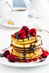 Stack of fried pancakes on a white plate. Chocolate, berries, raspberries, cranberries and blackberries on top. Gray concrete table, vintage forks, cup of tea with brown sugar, teapot.
