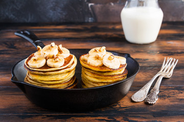 Stack of homemade pancakes with banana, maple syrup and walnuts in black cast iron skillet. Vintage forks, glass jar with milk, wooden rustic table.