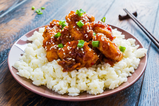 Crispy sesame chicken, chopped breast fillets, with a sticky sweet Asian sauce and white boiled rice on a plate on wooden table.
