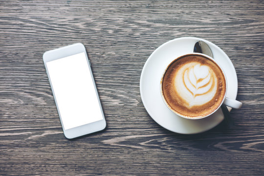 Mockup image of white mobile phone with blank white screen and hot latte coffee on vintage wood table in cafe