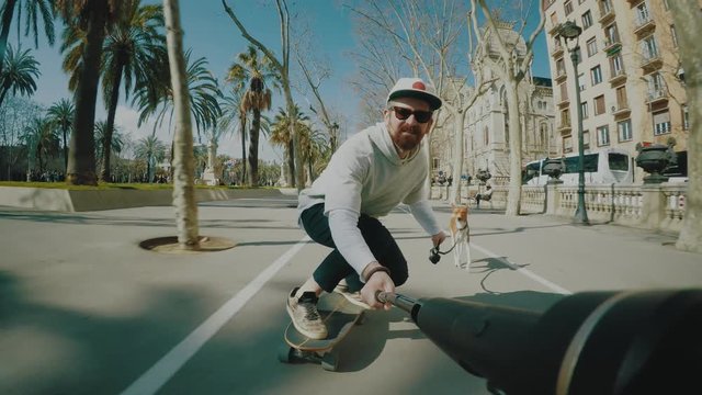 Happy bearded man rides his longboard on sunny street following his basenji dog on leash and making selfie on action camera on long gimbal stick ahead him.