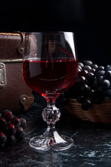 Glass of red wine on dark marble background. Cluster of blue grapes on on back background.