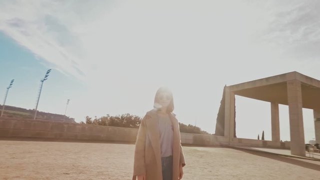 Camera turns around attractive girl in coat, smiling and posing in empty park with modern architecture at sunny winter day in spain