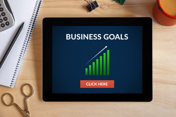 Business goals concept on tablet screen with office objects on wooden desk. All screen content is designed by me. View from above.