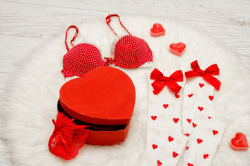 Fashion concept. Red box heart shaped with lace lingerie, white stockings with bows, red bra, red heart shaped candles on a white fur.