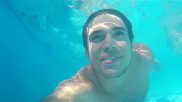 Handsome young man swimming in pool looking at camera smiling, underwater shot